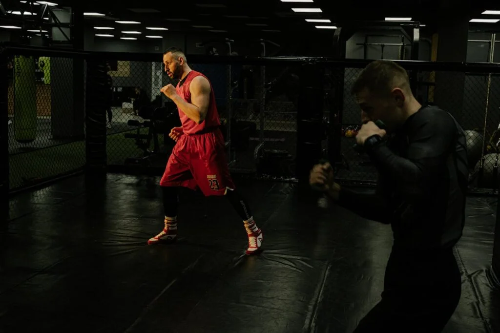 Two men practice boxing techniques in a dimly-lit gym, focusing on form and agility.